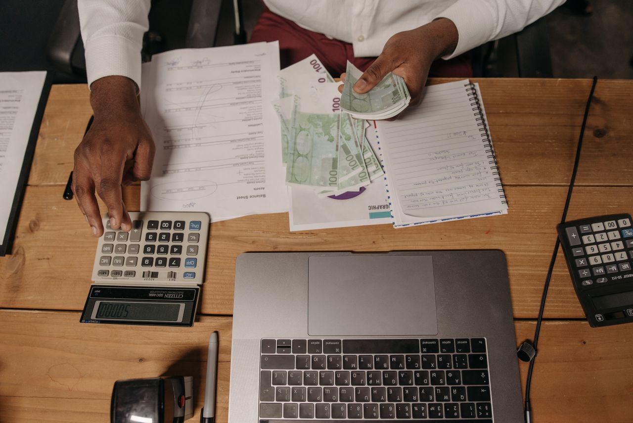 brand-01 Close-up of a person counting cash with documents and a laptop in an office setting.